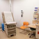 A clinical examination room with a brown examination table, orange chair, medical equipment on the wall, and a cabinet holding medical supplies.