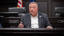 A man identified as Aging Secretary Jason Kavulich sits at a desk in a dark wood-paneled government hearing room.