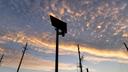 Silhouetted Flock Safety license plate reader against an orange-and-blue sunset sky, with utility poles and power lines below.