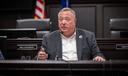 A man identified as Aging Secretary Jason Kavulich sits at a desk in a dark wood-paneled government hearing room.