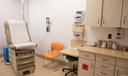 A clinical examination room with a brown examination table, orange chair, medical equipment on the wall, and a cabinet holding medical supplies.