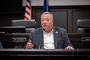A man identified as Aging Secretary Jason Kavulich sits at a desk in a dark wood-paneled government hearing room.