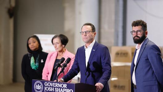 News conference featuring Governor Josh Shapiro at a podium, flanked by three individuals in a spacious industrial setting with cardboard boxes in the background. News conference featuring Governor Josh Shapiro at a podium, flanked by three individuals in a spacious industrial setting with cardboard boxes in the background.