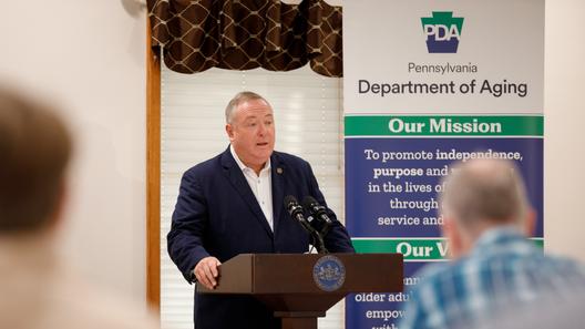 Pennsylvania Department of Aging Secretary Jason Kavulich speaks at a podium during an event, with a banner behind him outlining the department's mission to support older adults.
