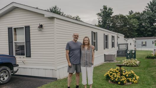 A man and woman stand in front of a mobile home with yellow flowers next to them. A man and woman stand in front of a mobile home with yellow flowers next to them.