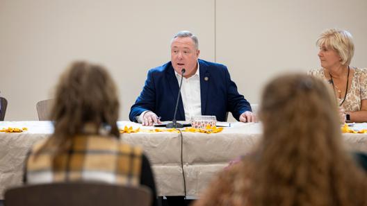 Aging Secretary Jason Kavulich speaks into a microphone during a meeting, with attendees in the foreground and a woman seated beside him.