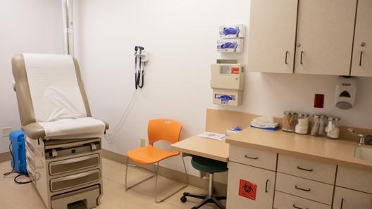 A clinical examination room with a brown examination table, orange chair, medical equipment on the wall, and a cabinet holding medical supplies.