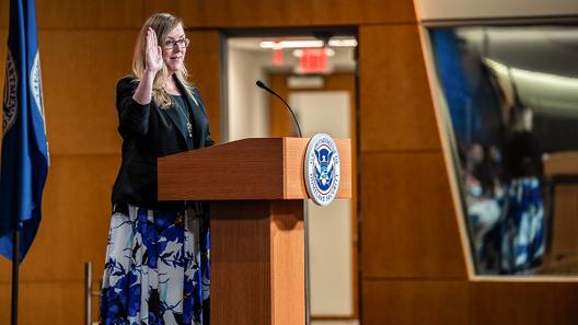 Jennifer Higgins, then-USCIS deputy director, swears in new citizens while raising her hand at a podium with the USCIS seal.