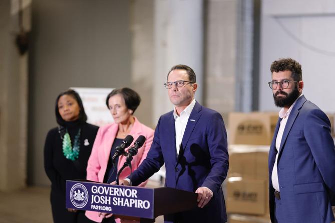 News conference featuring Governor Josh Shapiro at a podium, flanked by three individuals in a spacious industrial setting with cardboard boxes in the background.