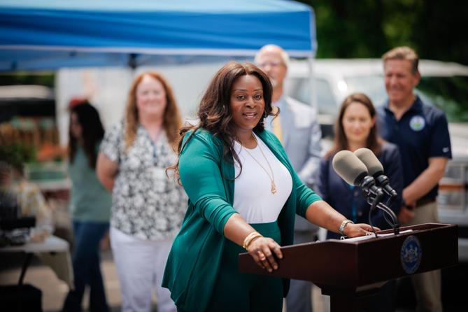 Sally Zubairu-Cofield speaks at an outdoor news conference from behind a podium with two microphones, with several people and a blue canopy behind her. Sally Zubairu-Cofield speaks at an outdoor news conference from behind a podium with two microphones, with several people and a blue canopy behind her.