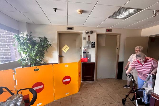 Two women, one leaning on a walker, stand in a hall near two elevators. One elevator with an out of order sign on the door has four barriers in front of it.