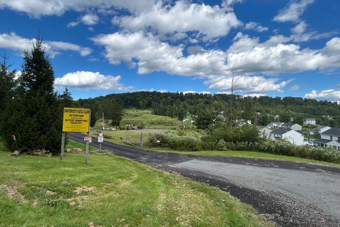 A yellow sign next to a driveway says “University Rifle Club. Attention this is an active shooting facility.” To the left of the road is a housing development and a hill in the background. A yellow sign next to a driveway says “University Rifle Club. Attention this is an active shooting facility.” To the left of the road is a housing development and a hill in the background.