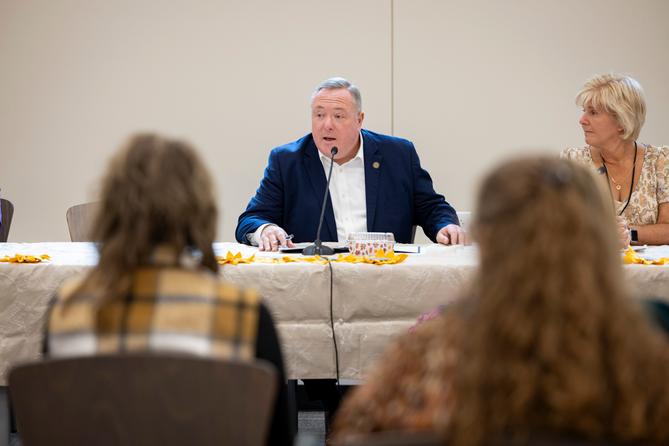 Aging Secretary Jason Kavulich speaks into a microphone during a meeting, with attendees in the foreground and a woman seated beside him. Aging Secretary Jason Kavulich speaks into a microphone during a meeting, with attendees in the foreground and a woman seated beside him.