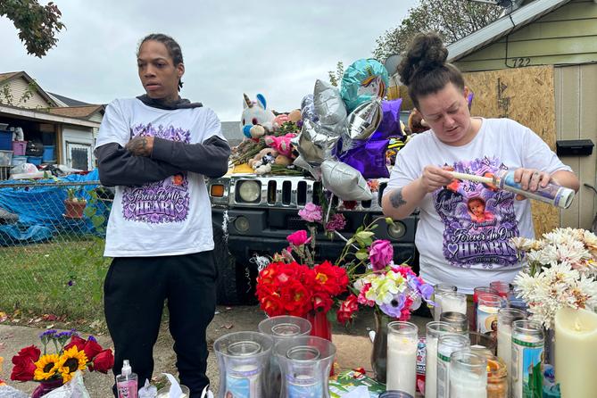 A man and woman stand at a memorial of flowers, balloons, and candles.